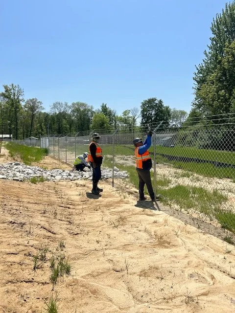 Two crew members installing chain link fence along a sandy embankment